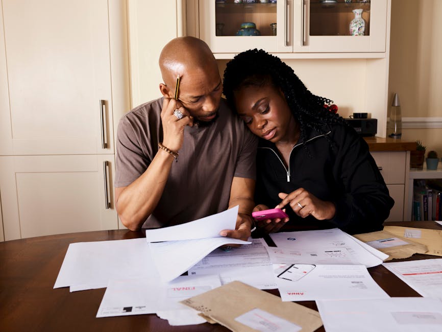 A couple sits at a table managing domestic finances, evaluating documents and using a smartphone.