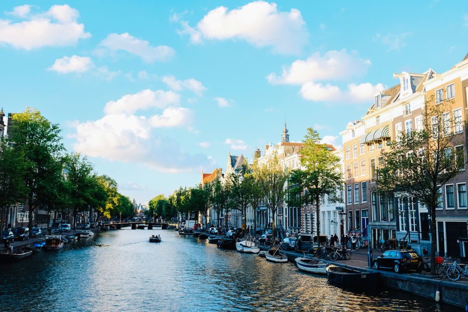 Magnificent cityscape of Amsterdam with traditional boats moored on calm river near historic buildings under cloudy blue sky