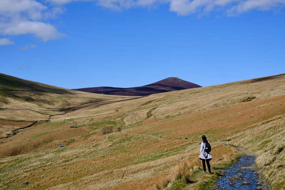 A woman with a backpack stands in the scenic hills of Threlkeld, England, under a clear blue sky.