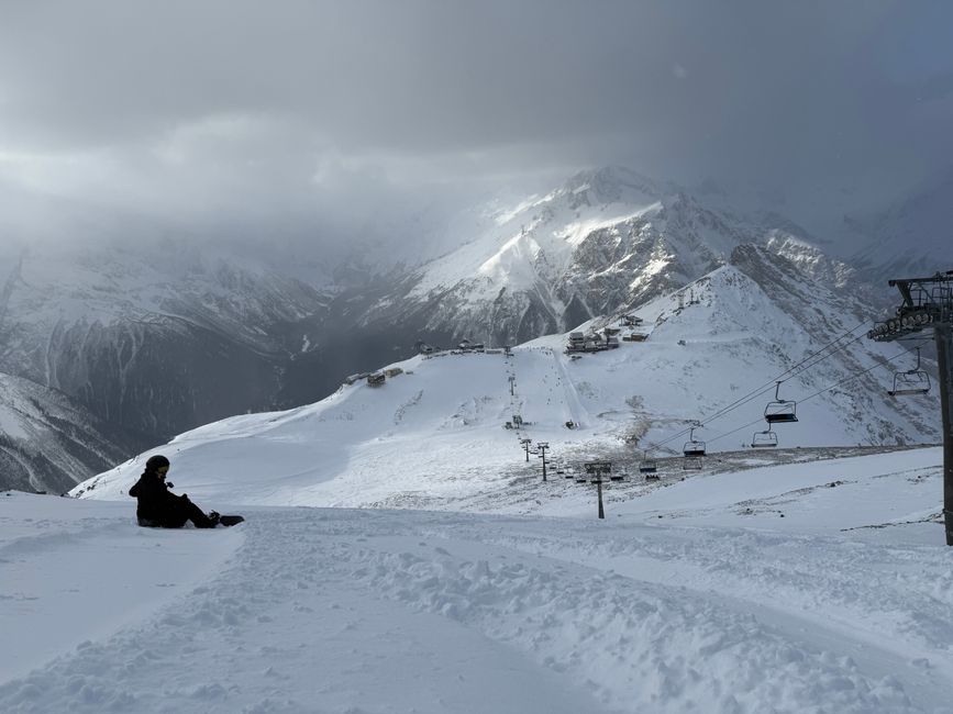 A lone snowboarder sits on a snow-covered mountain with ski lifts in view.