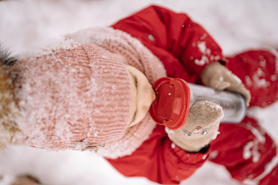A child in snow-proof clothes enjoys a drink from a tumbler in a snowy outdoor setting.