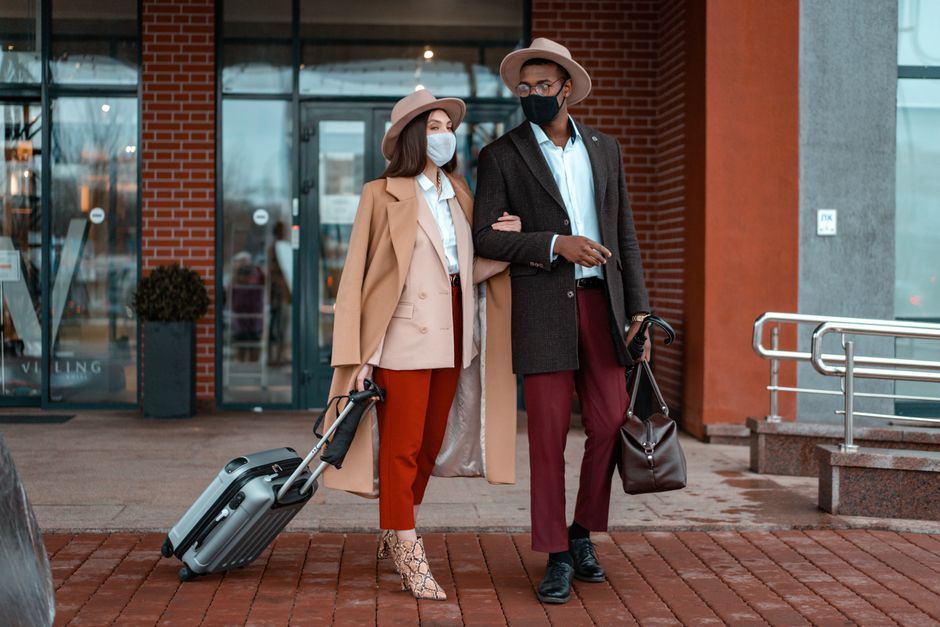 A fashionable couple, wearing face masks, stands with luggage outdoors, ready to travel.