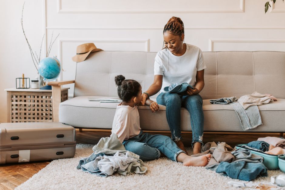 African American mother and daughter packing suitcase on sofa, smiling and enjoying family time.